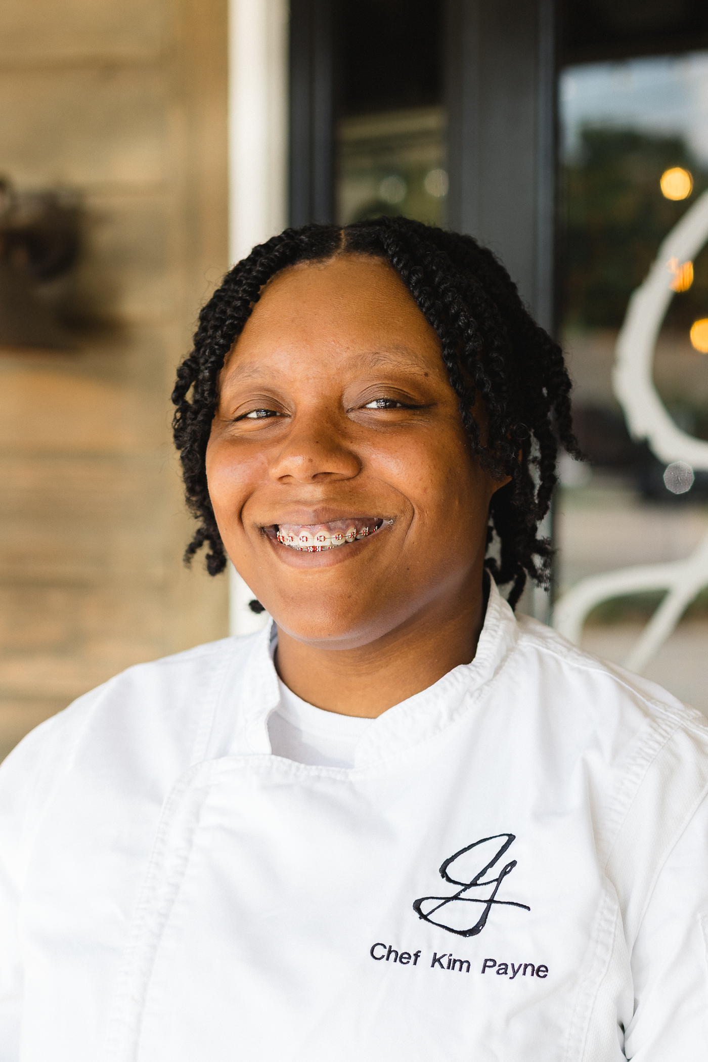 Smiling Black female chef in a white coat with 'Chef Kim Payne' embroidered on the chest.